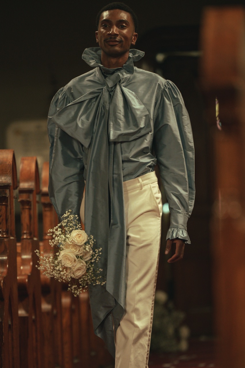 Model walks down the aisle carrying a bouquet of white roses and baby's breath during Tanner Fletcher's immersive bridal fashion show at New York Bridal Fashion Week, wearing a dramatic slate-blue bow blouse with ivory tailored trousers inside a historic church.